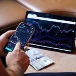 A person using a smartphone and tablet for stock market analysis, with graphs and cash visible.