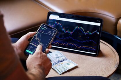 A person using a smartphone and tablet for stock market analysis, with graphs and cash visible.