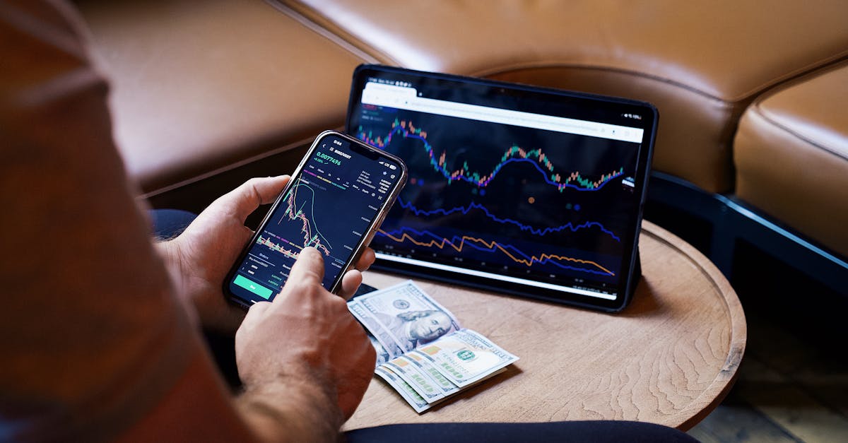 A person using a smartphone and tablet for stock market analysis, with graphs and cash visible.